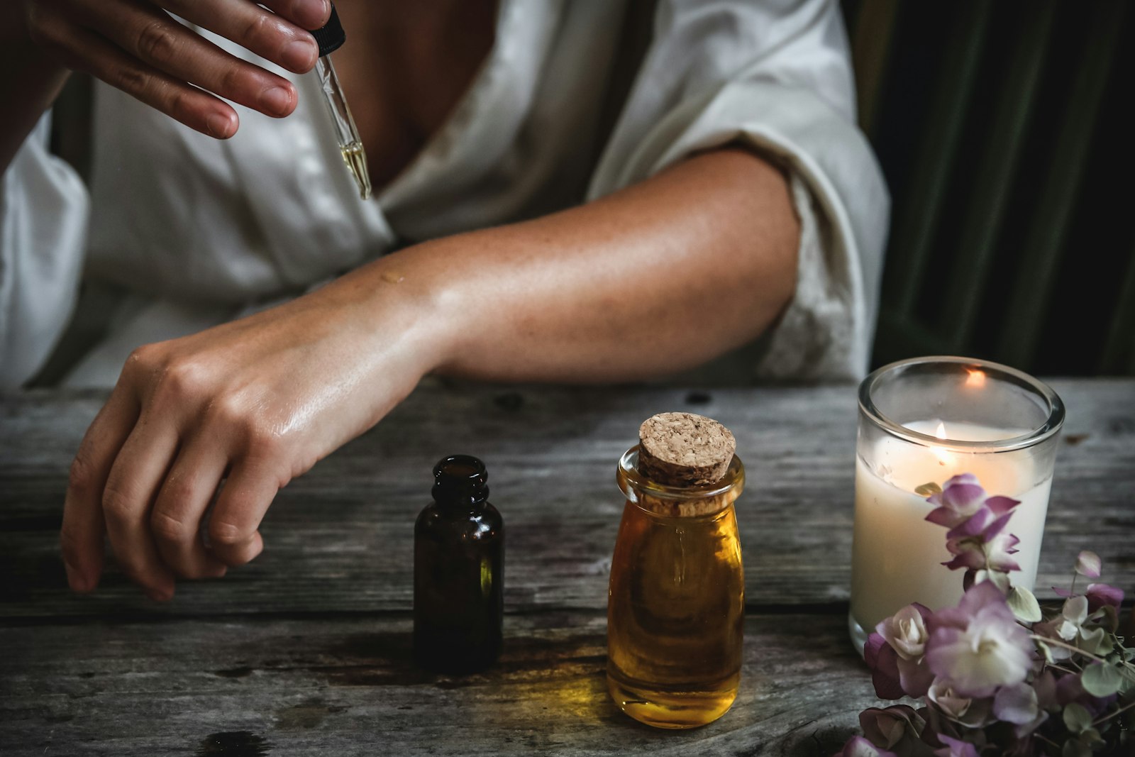 Hands applying essential oil beside a lit candle and fresh flowers